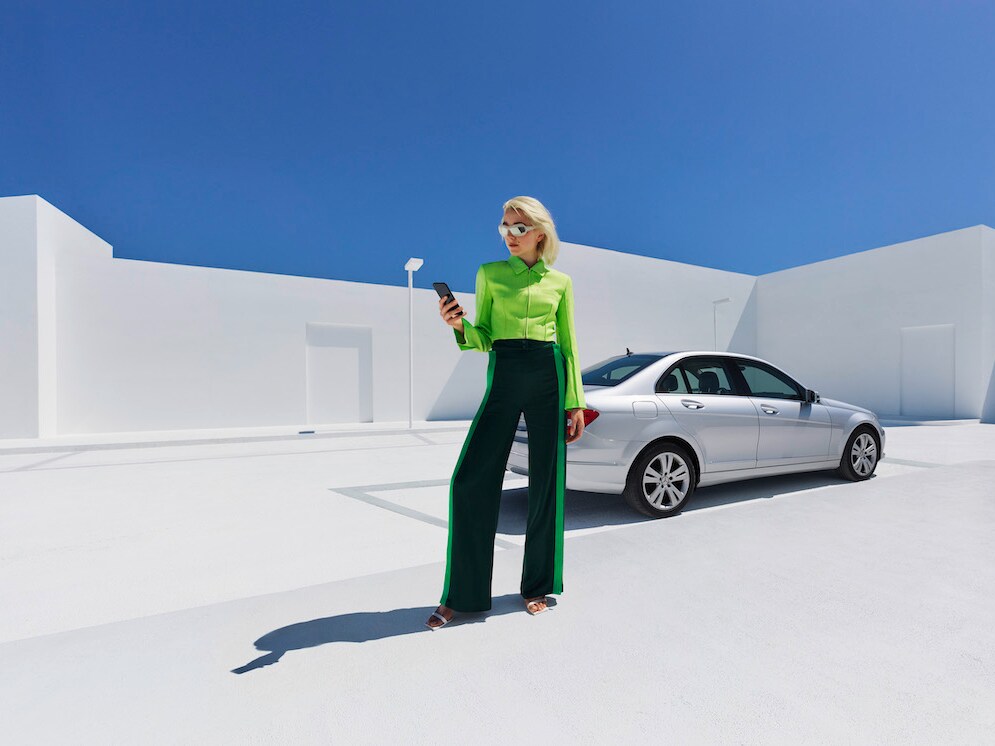 Eine Frau steht vor ihrem Mercedes-Benz und schaut auf ihr Smartphone.