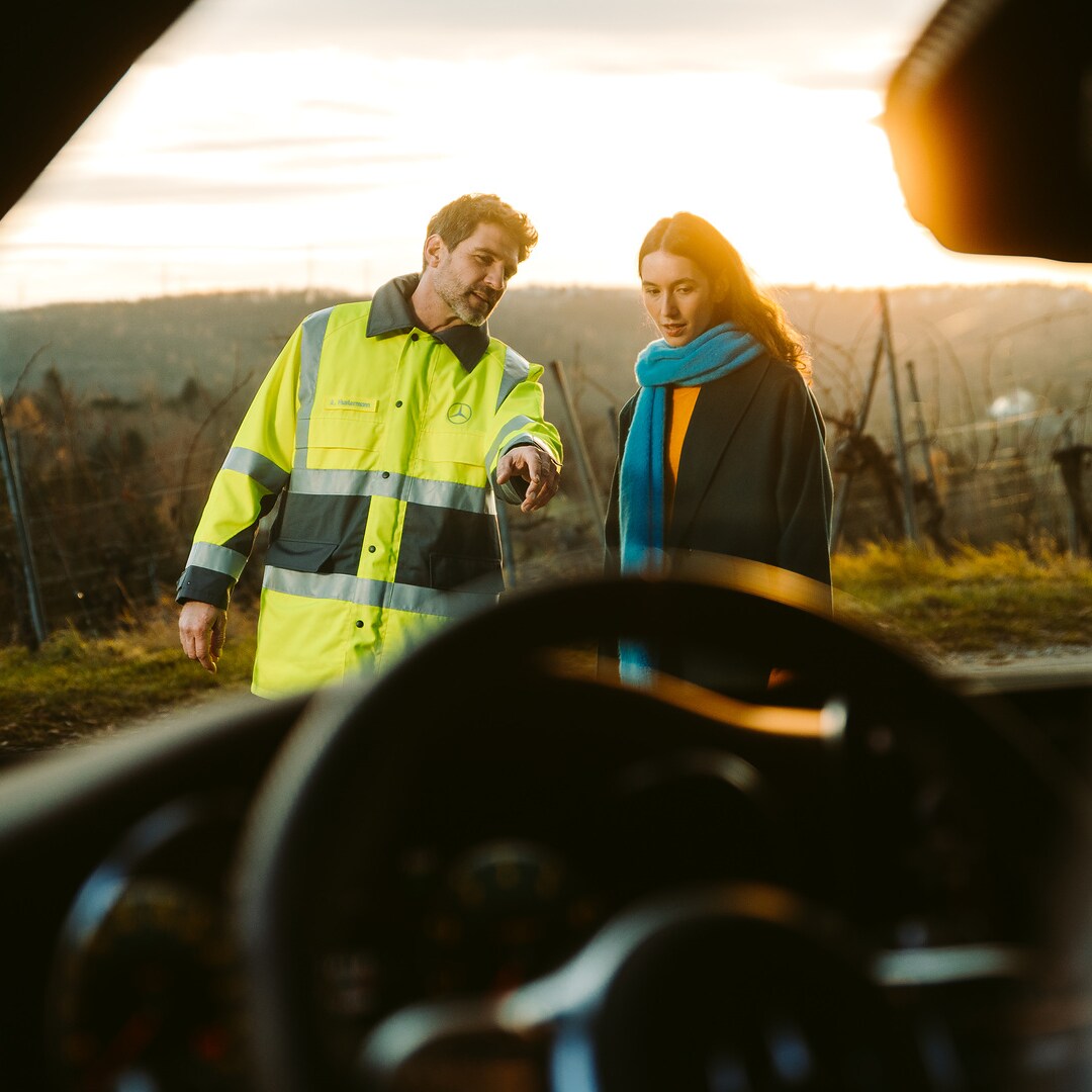 A travers le pare-brise d'une Mercedes, nous voyons un technicien de service qui se tient à côté de la conductrice devant le véhicule et le montre du doigt en guise d'explication.