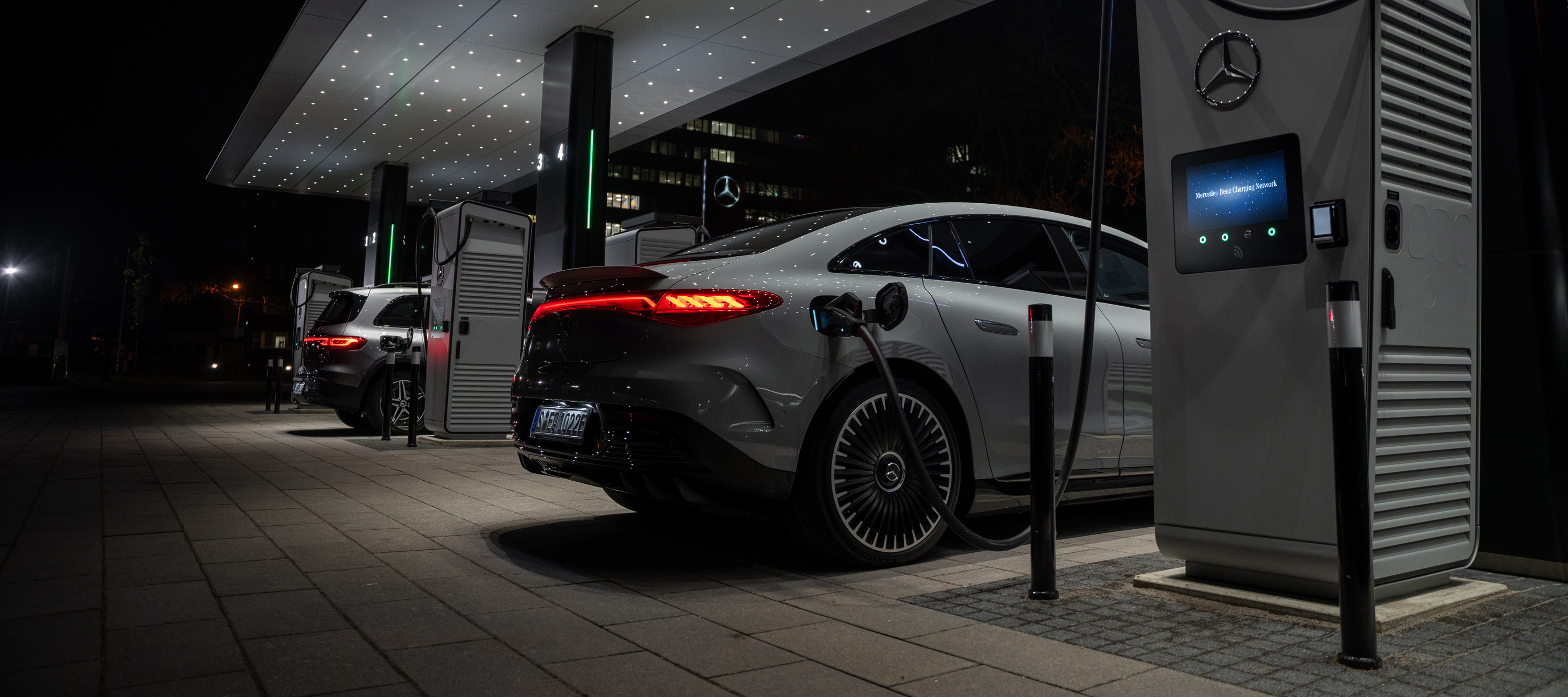 Mercedes-Benz Ladestation Das Bild zeigt eine nächtliche Szene, in der eine Mercedes-Benz EQS Limousine über ein Ladekabel mit einer Hochleistungsladestation verbunden ist.