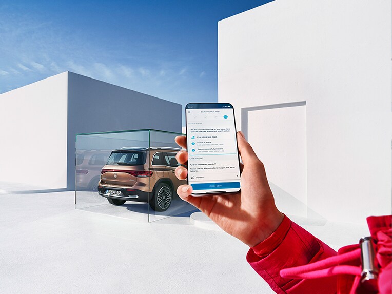 Une personne tient son téléphone portable dans la main et utilise l'application Mercedes me. Un EQB stationne en arrière-plan.