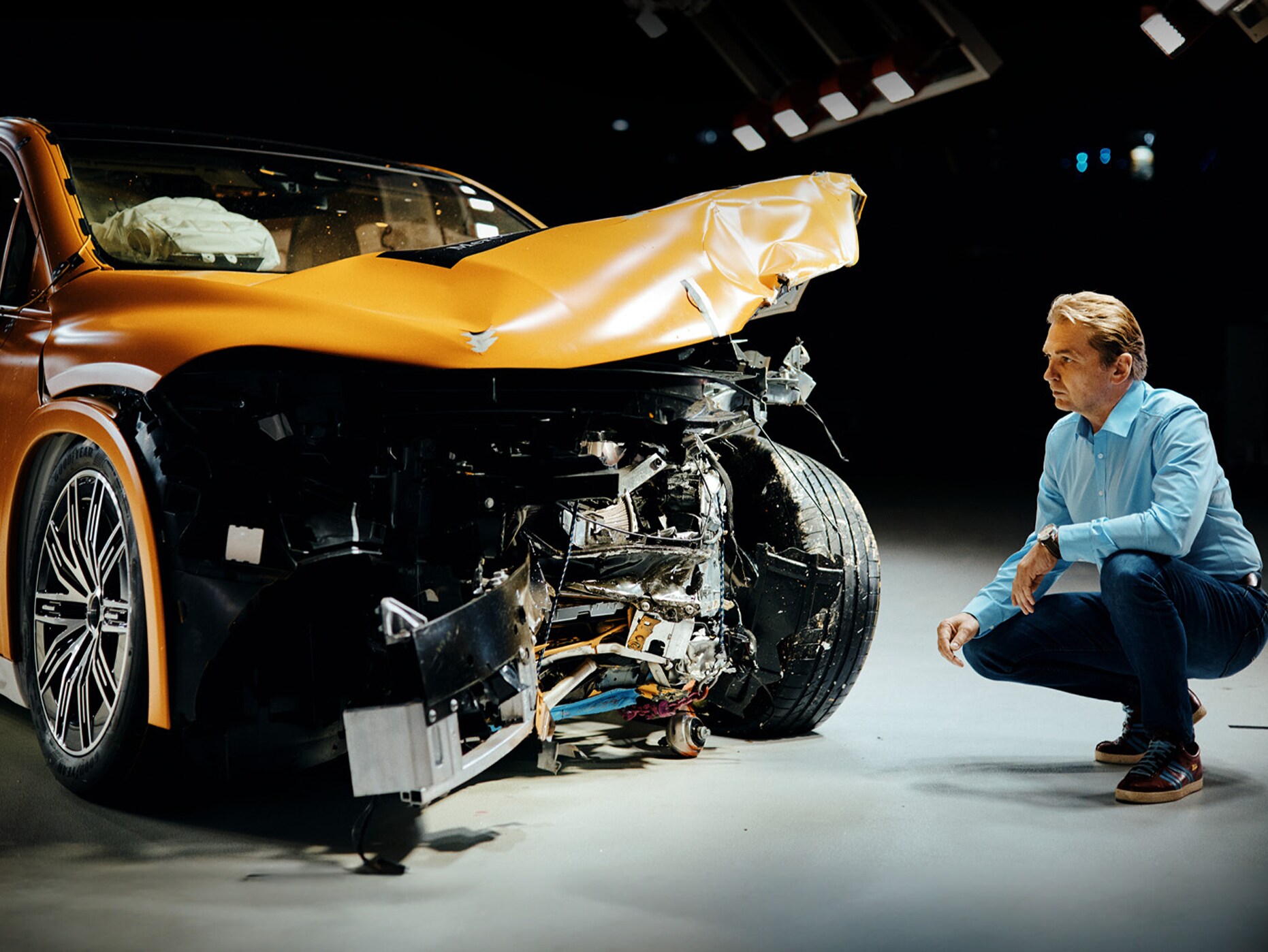 Un expert en essais de collision examine une voiture Mercedes-Benz accidentée à l'avant en s'accroupissant à côté d'elle.