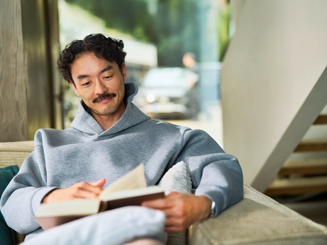 Client assis à l'intérieur en train de lire un livre. Derrière lui, on aperçoit à travers une fenêtre une voiture en cours d'entretien par un spécialiste.