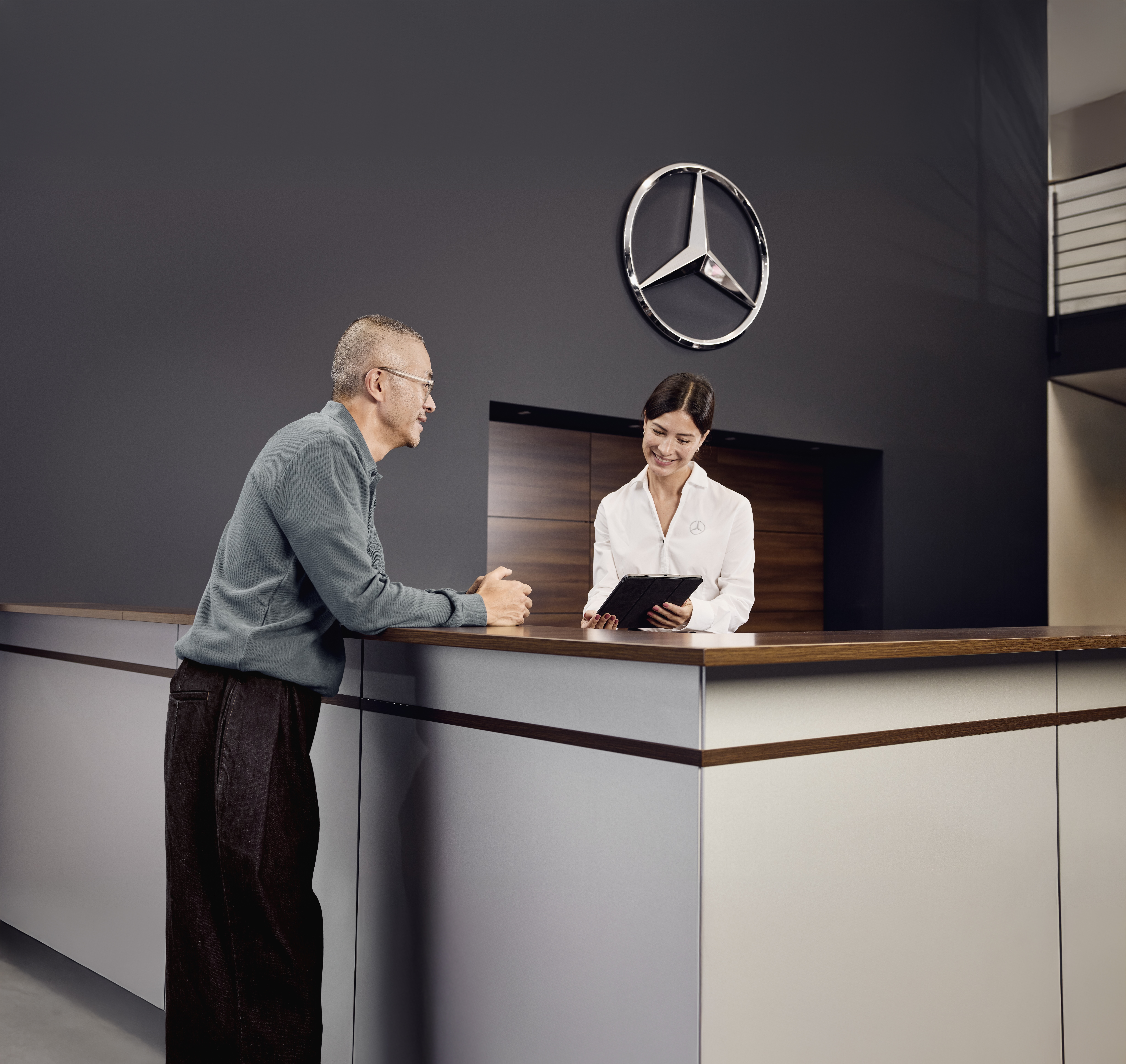 Beratung zu Mercedes-Benz Originalteilen Empfangsbereich der Serviceabteilung eines Mercedes-Benz-Händlers, wo ein Kunde mit einem Serviceberater am Empfang die Einzelheiten der Fahrzeugwartung bespricht.