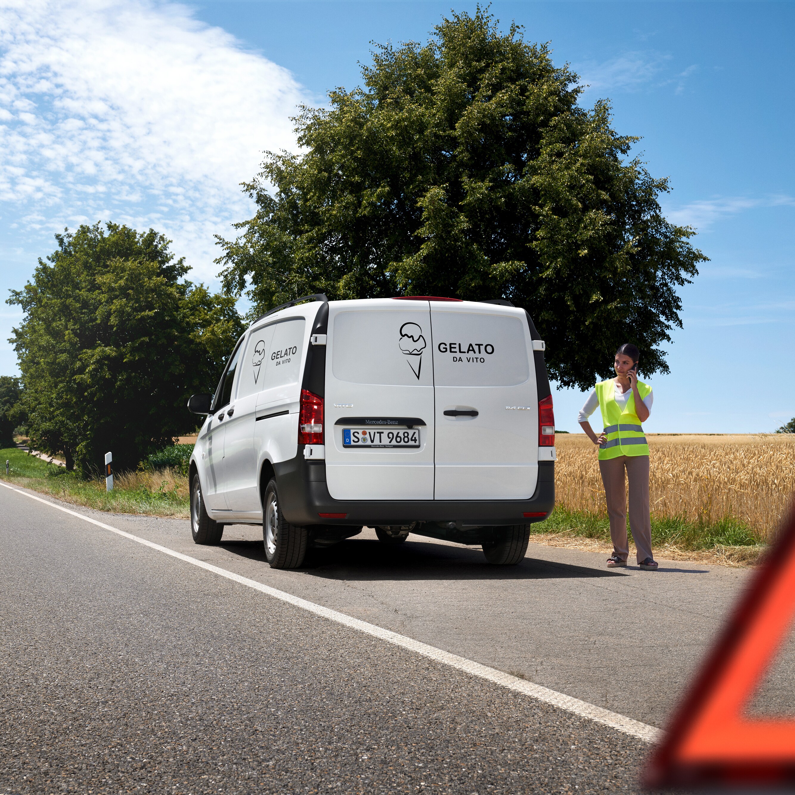 Eine Frau ruft die Mercedes-Benz Service24h-Hotline an; ein Mercedes-Benz Sprinter steht am Rand einer Landstraße.