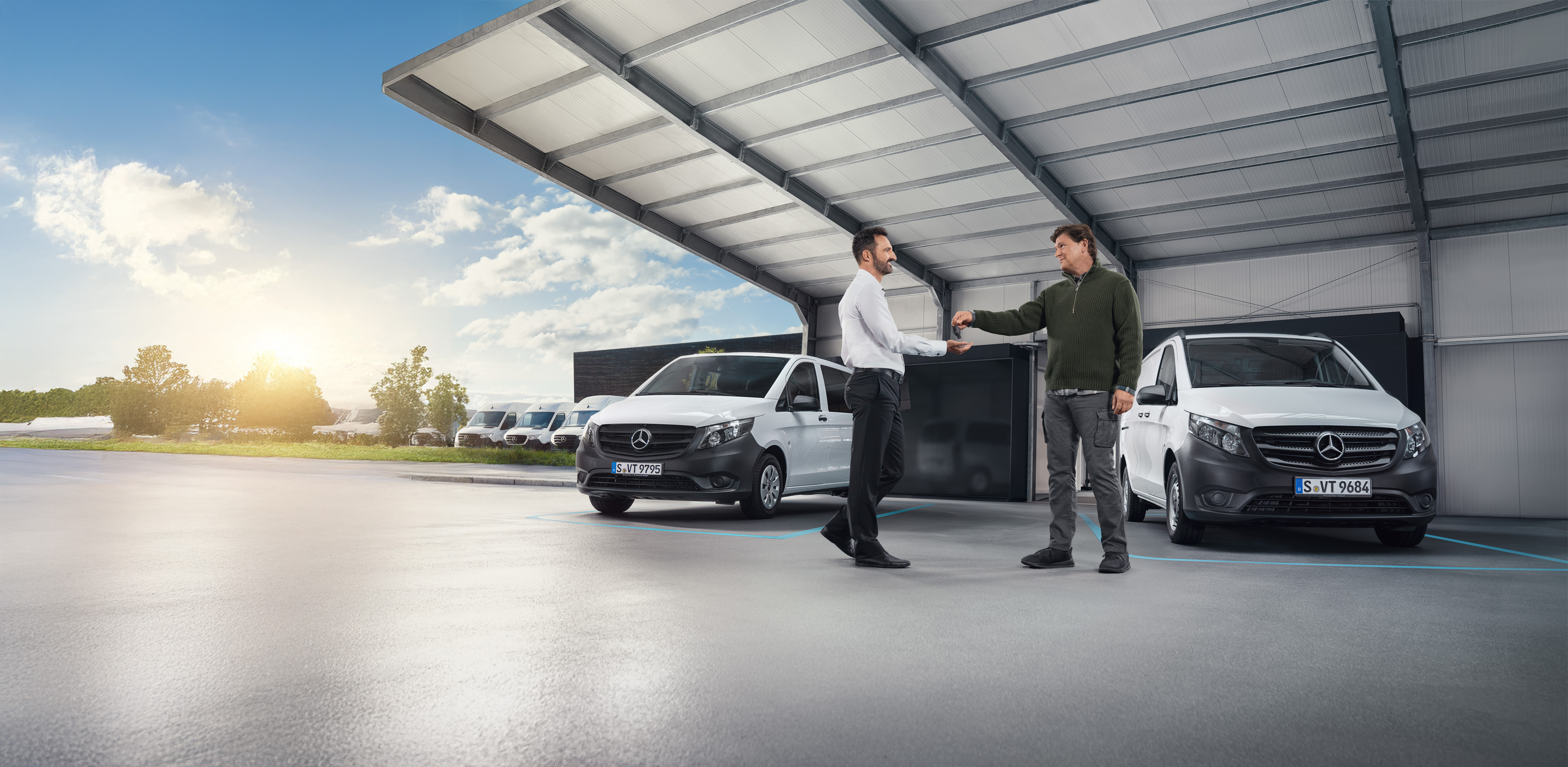 Mercedes-Benz | Transporterbetreuung | Individuelle Unterstützung Zwei Männer unterhalten sich vor einer Reihe weißer Mercedes-Benz Transporter in einer Halle