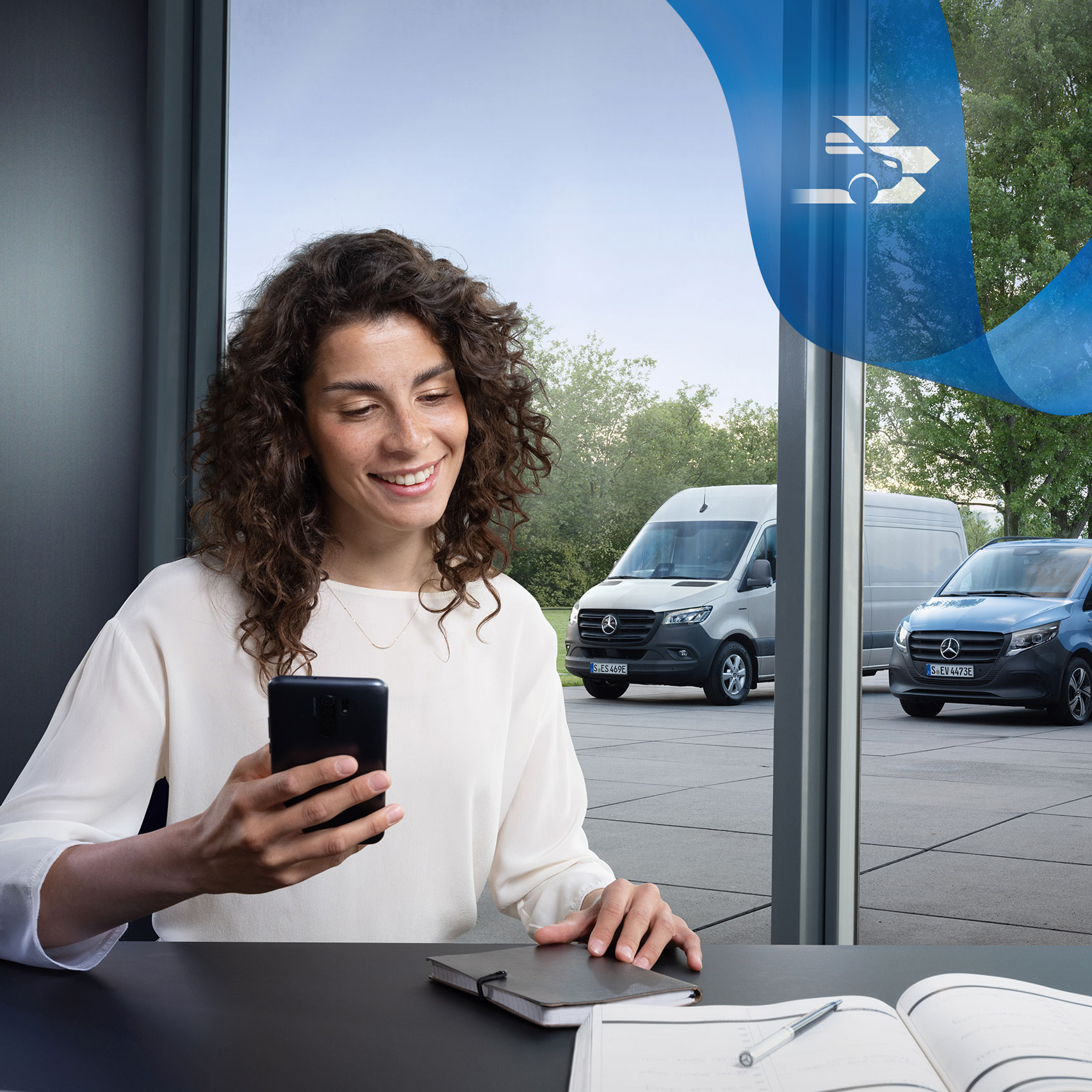 Mercedes-Benz | Assistance pour utilitaires légers | Centre d'assistance à la clientèle Femme assise dans un bureau avec un smartphone et une série d’utilitaires légers Mercedes-Benz en arrière-plan.