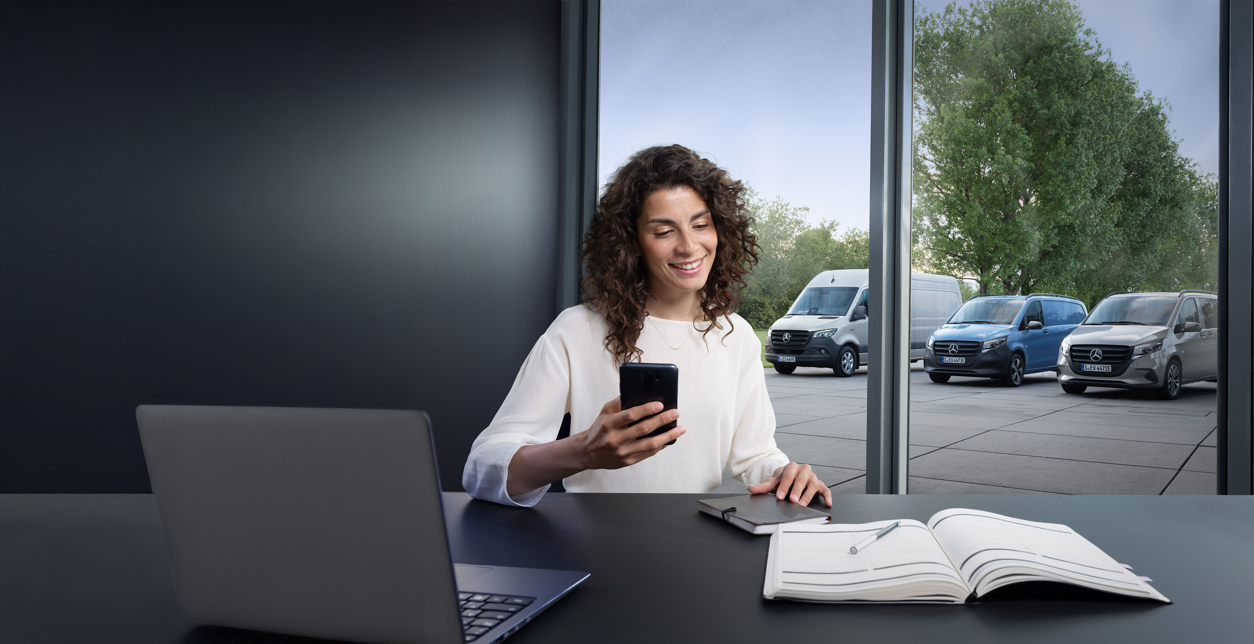 Mercedes-Benz | Confort | Point de service agréé Mercedes-Benz Femme recherchant un partenaire de service agréé Mercedes-Benz sur son smartphone, avec plusieurs utilitaires Mercedes-Benz en arrière-plan.