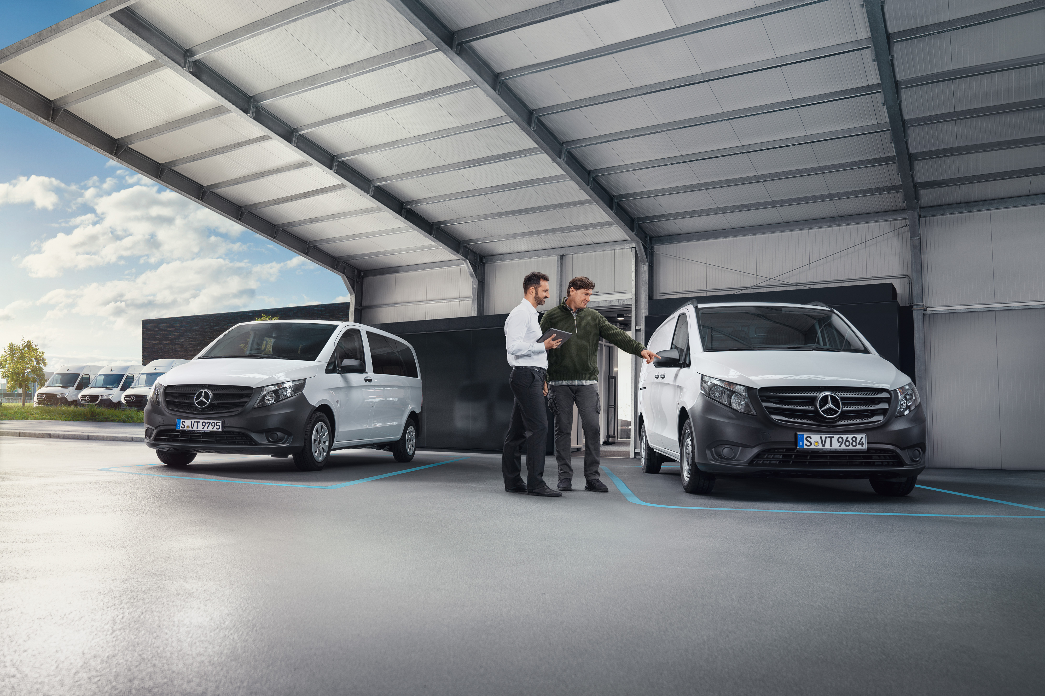 Mercedes-Benz | Confort | Accueil numérique des clients Deux personnes discutant dans un atelier devant deux utilitaires légers Mercedes-Benz blancs.