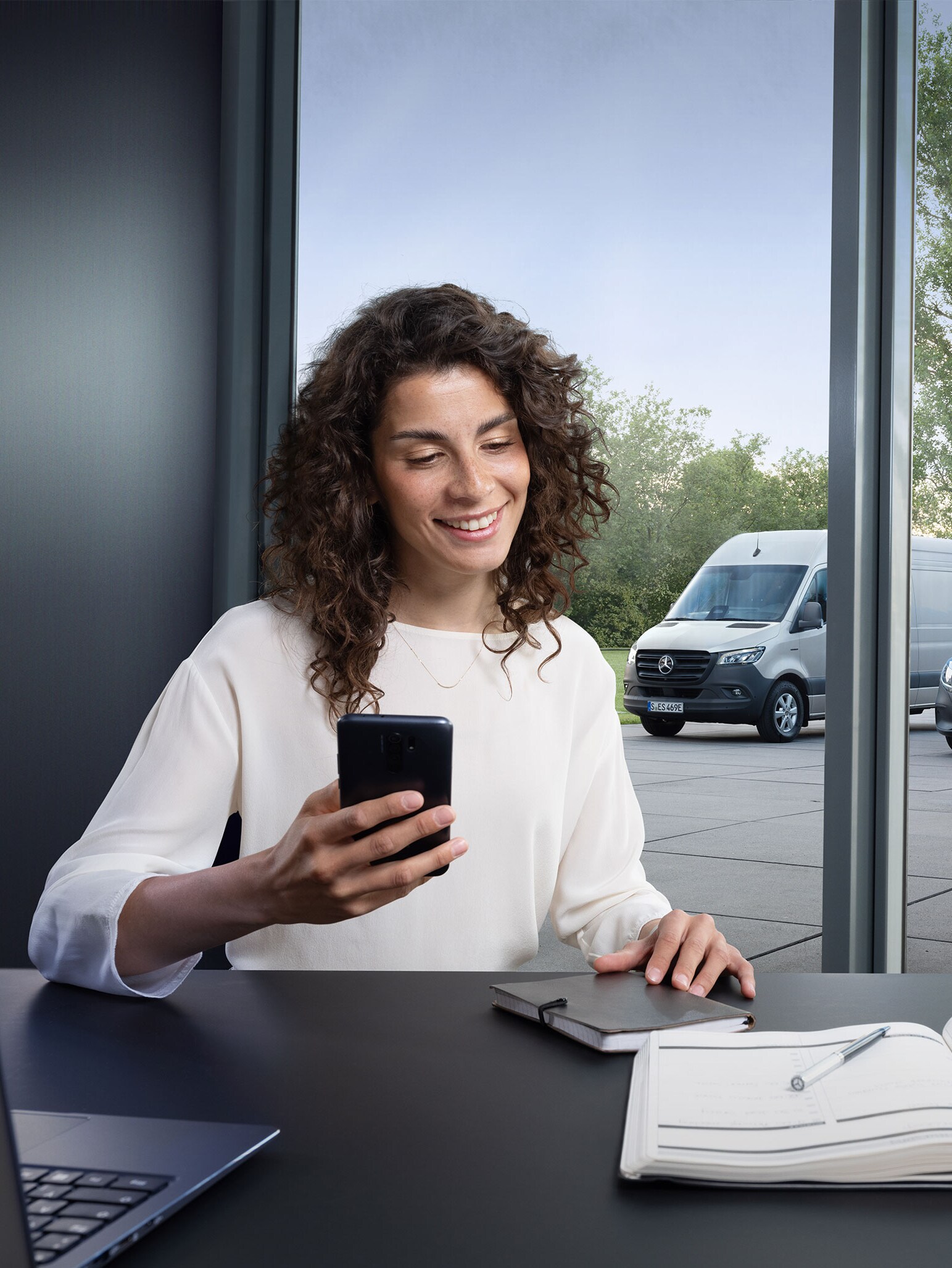 Femme réservant un service par smartphone avec une gamme d’utilitaires léger Mercedes-Benz en arrière-plan.
