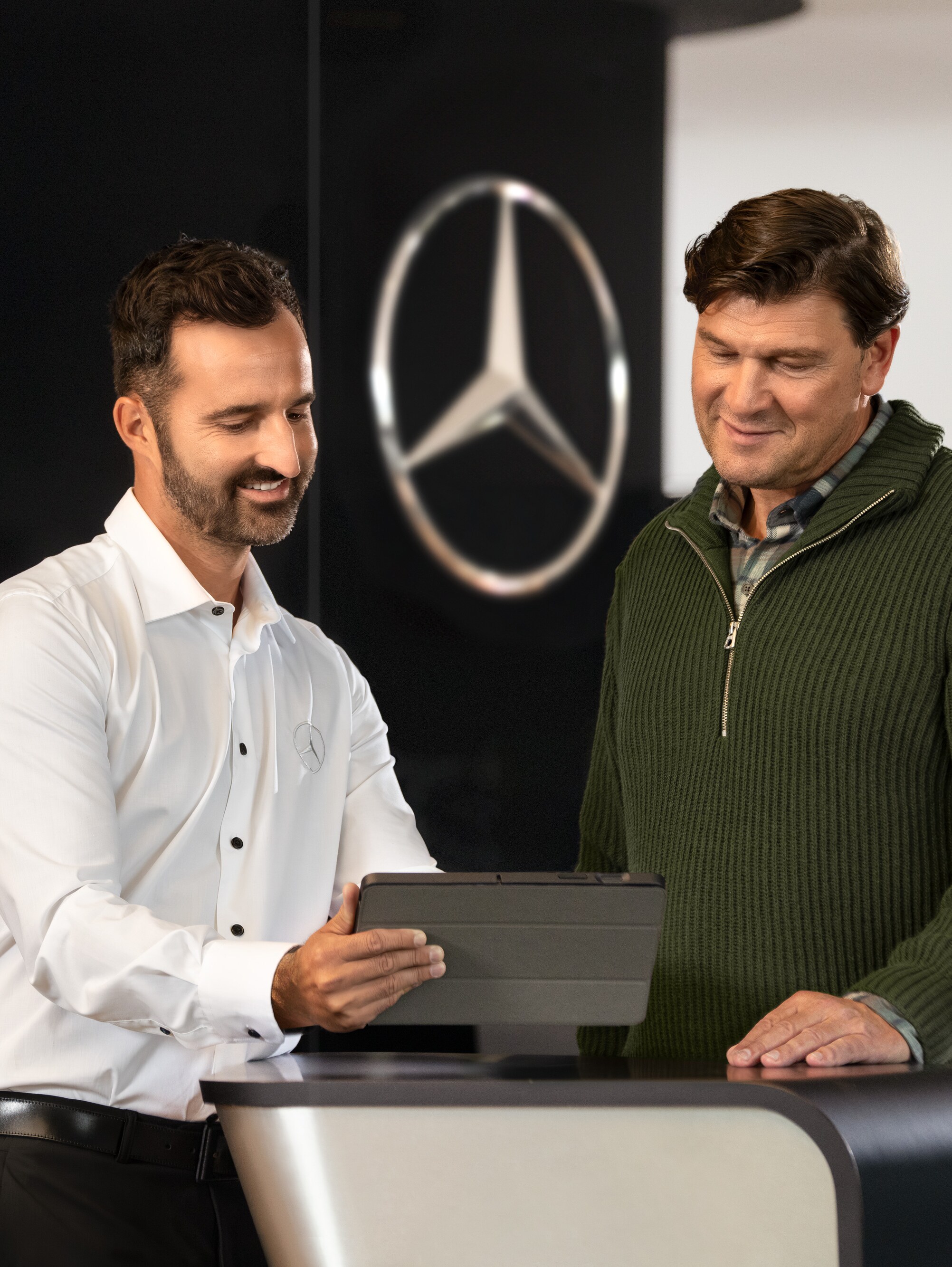 Zwei Männer unterhalten sich an einem Tresen vor dem Mercedes-Benz Stern, während sie auf ein Tablet schauen.