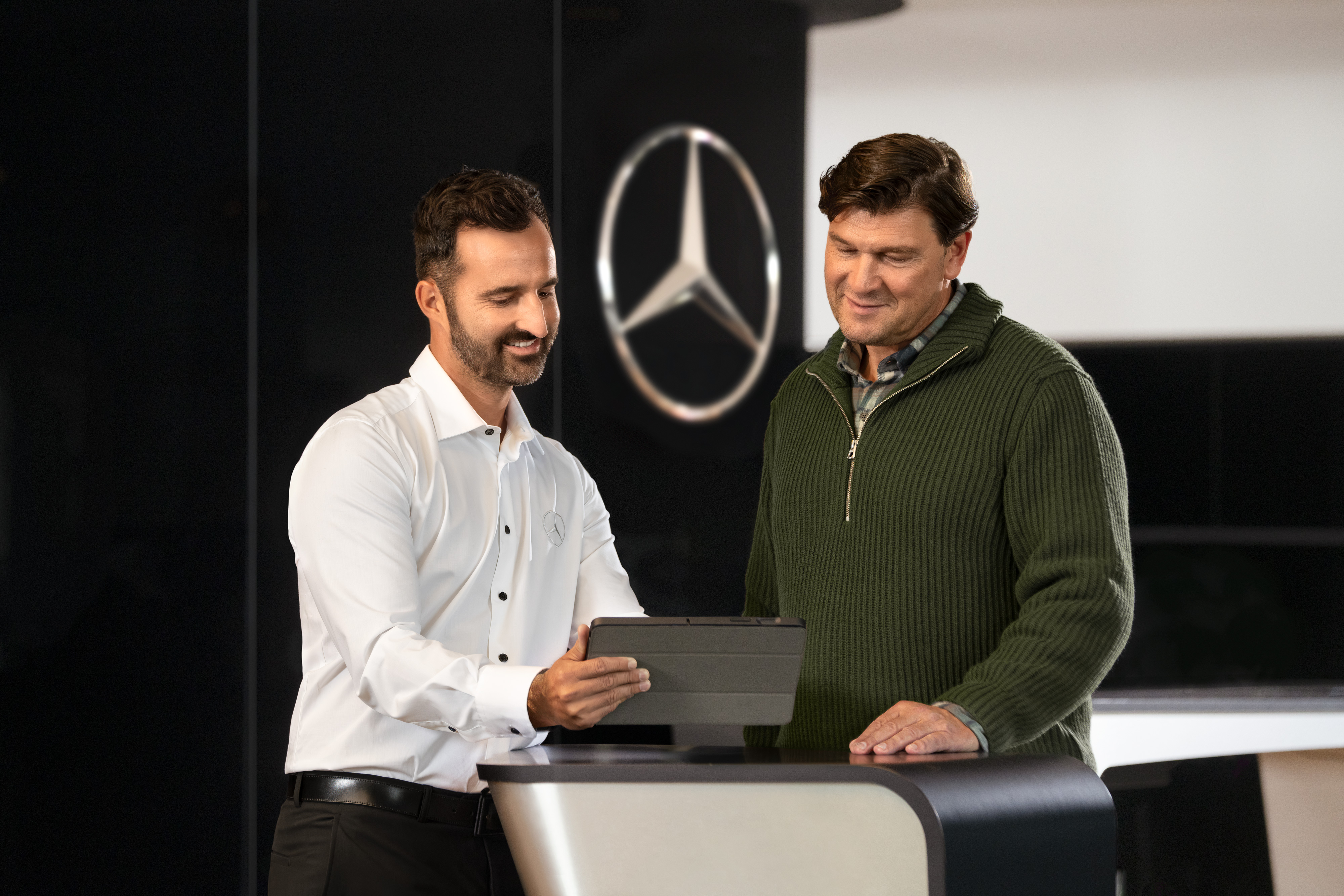 Mercedes-Benz | Dienstleistungen im Überblick Zwei Männer unterhalten sich an einem Tresen vor dem Mercedes-Benz Stern, während sie auf ein Tablet schauen.