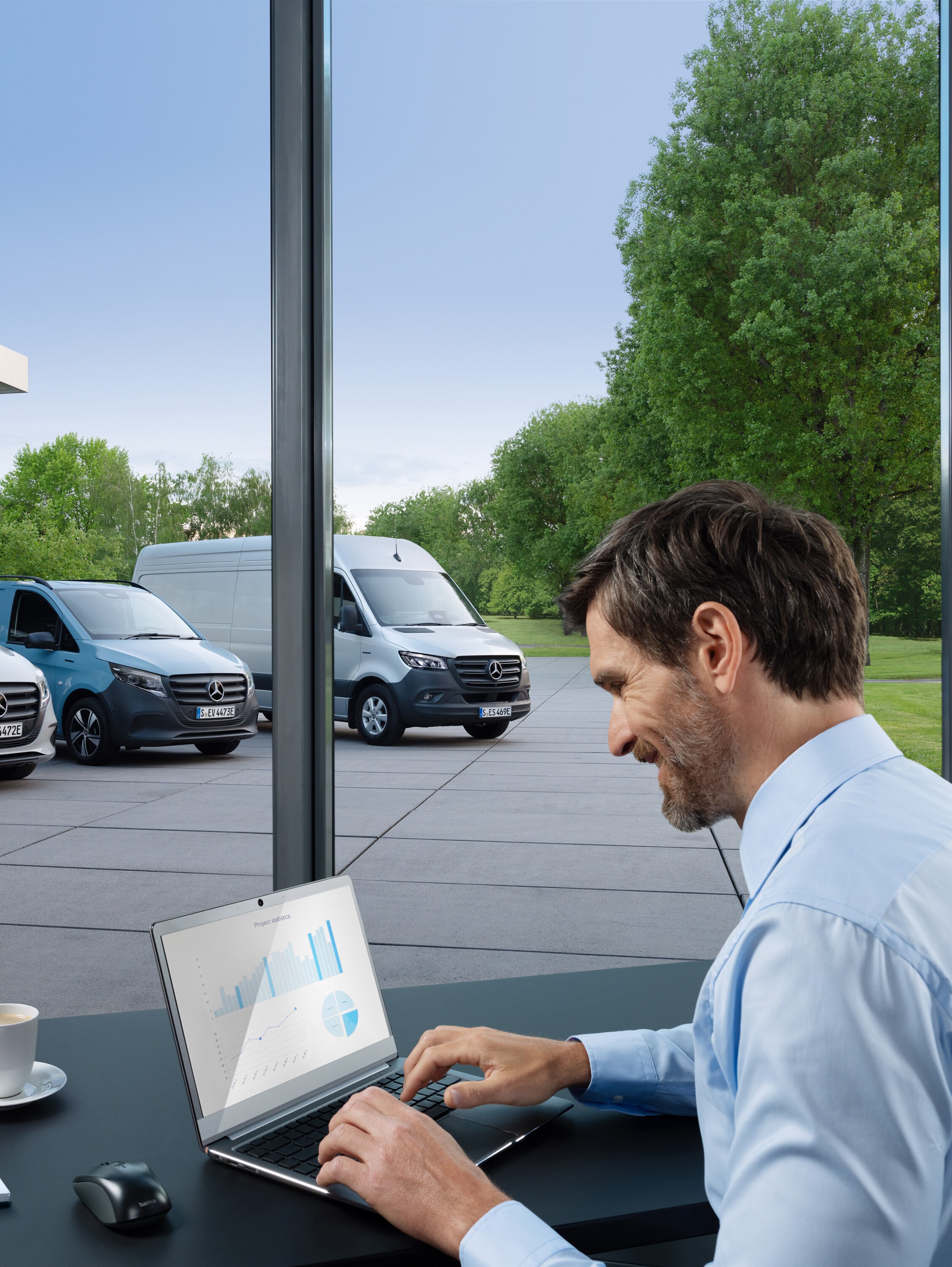 Ein Mann arbeitet an einem Laptop, draußen steht eine Reihe von Mercedes-Benz Transportern.