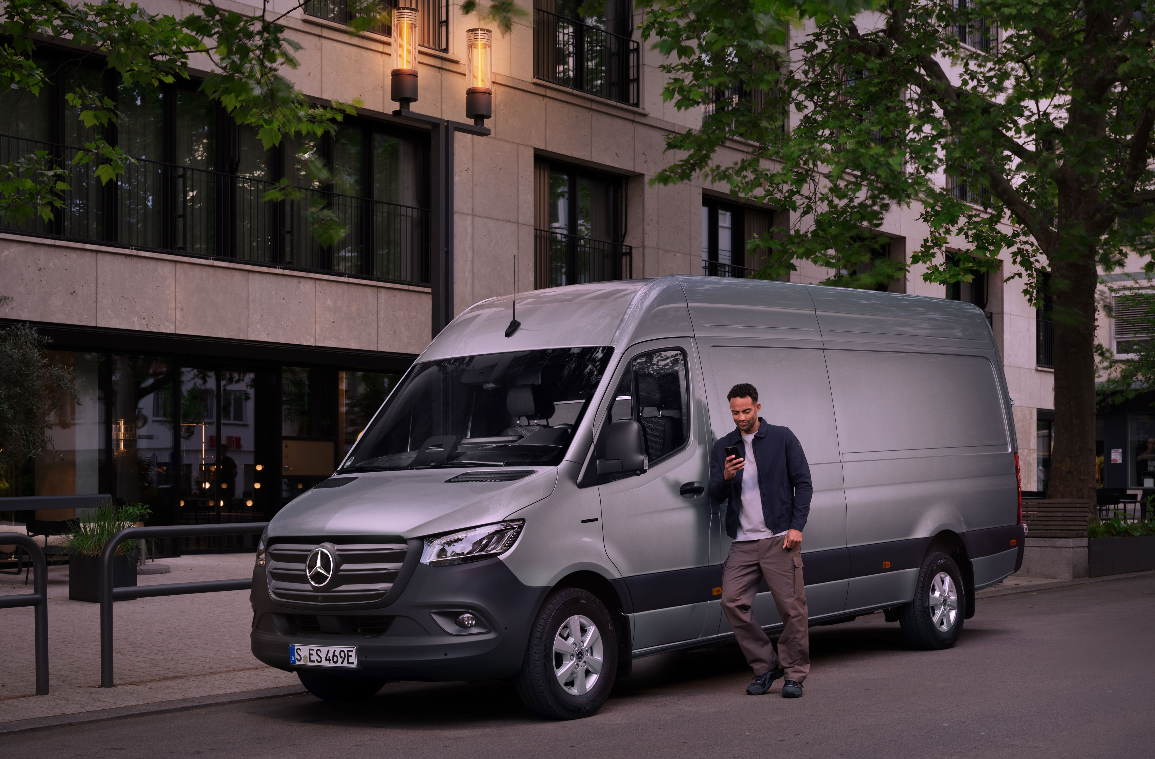 Mercedes-Benz | Réparation | Digital Damage Notification Un homme s'appuie sur un Mercedes-Benz Sprinter couleur argent et remplit une déclaration de dommage sur son téléphone portable.
