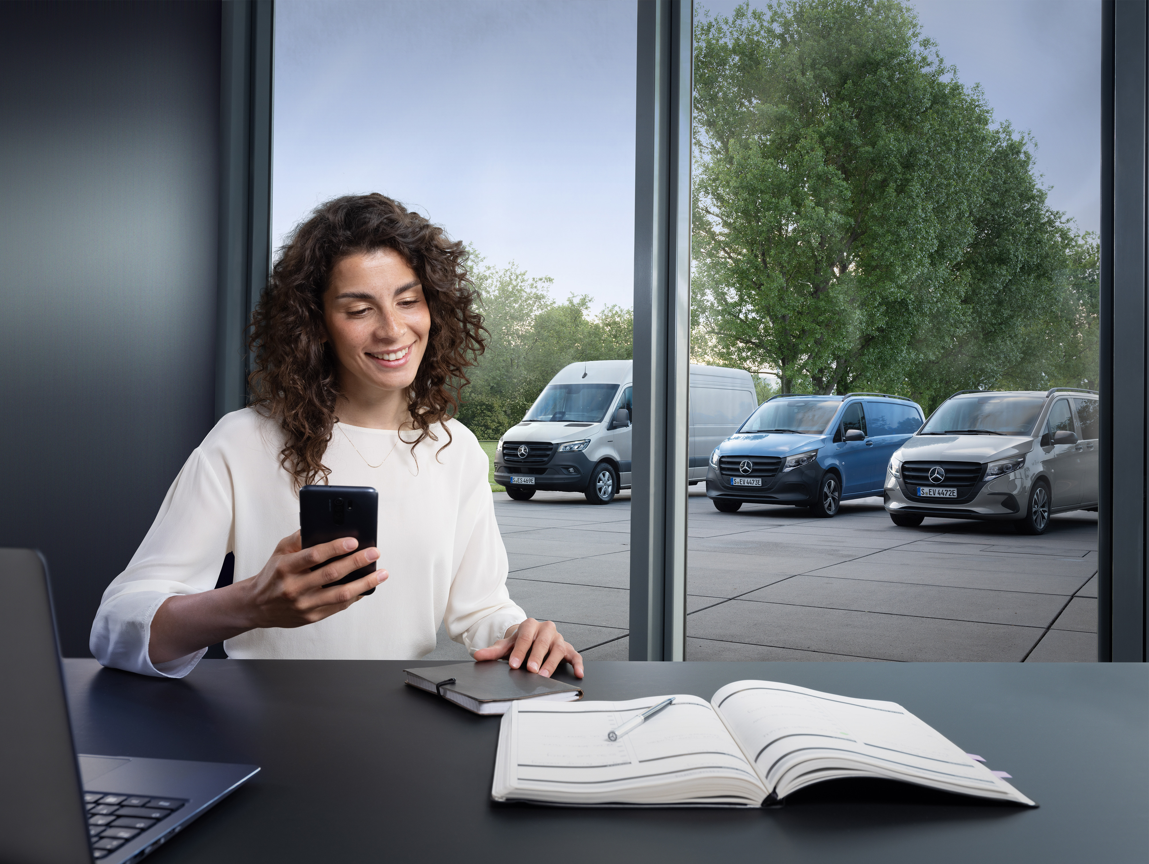 Mercedes-Benz | Van ServiceCare | Prise de rendez-vous Une femme planifie sur son smartphone devant une gamme d’utilitaires Mercedes-Benz en arrière-plan.