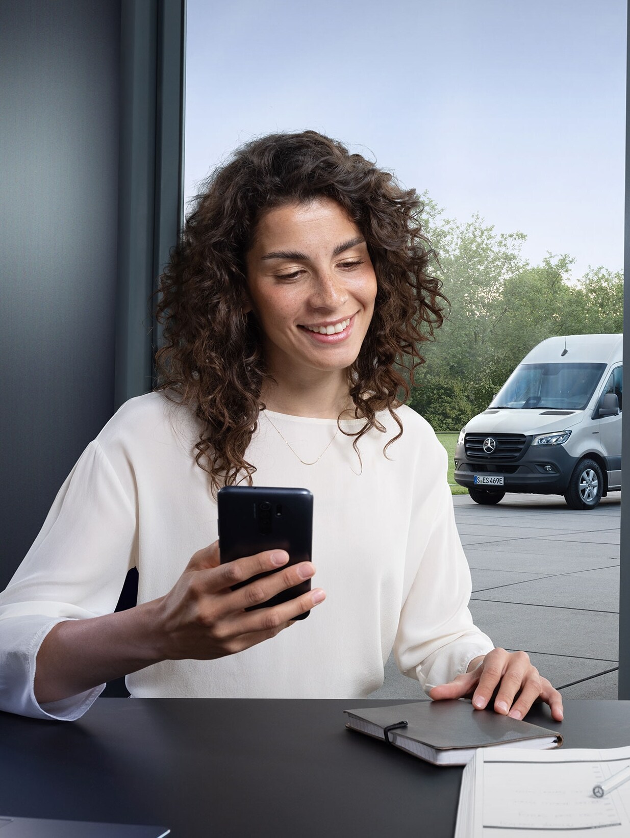 Frau, die mit einem Smartphone die Fuhrparküberwachung mehrerer Mercedes-Benz Transporter am Bürofenster steuert.